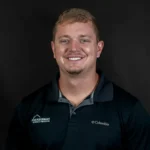 Professional headshot of smiling man in dark Columbia polo shirt against gray background